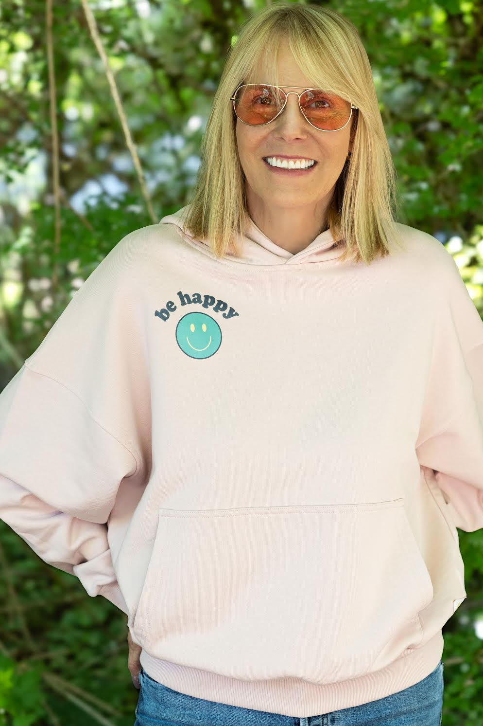 Woman wearing a light pink hoodie with a logo and text, standing outdoors with greenery in the background.